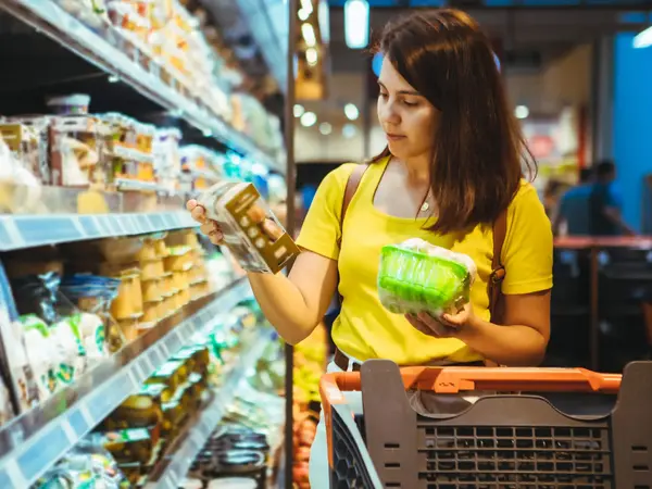 Eine Frau steht in einem Supermarkt vor einem Regal mit gekühlten Produkten und hält eine braune und grüne Lebensmittelverpackung in der einen Hand.