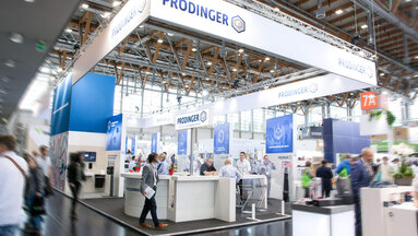 In einer Messehalle ist ein Stand von PRODINGER zu sehen, auf dem Besucher von Mitarbeitern beraten werden.